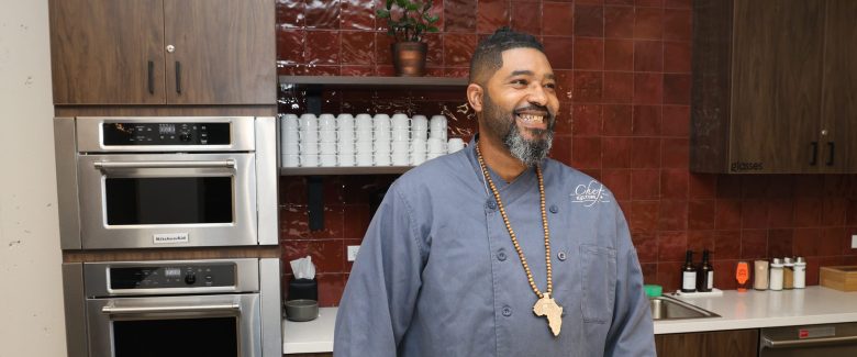 A Black man in a chef coat wearing an Africa necklace