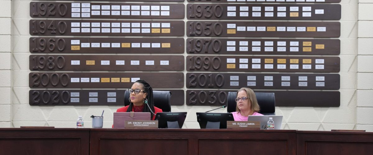 Two people sit at a school board meeting.